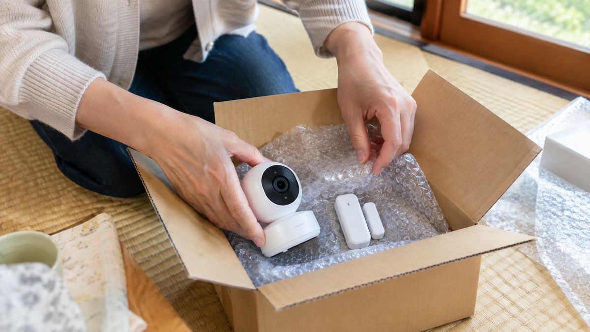 返却用の段ボール箱に、白い室内カメラと開閉センサーを緩衝材で包んでしまう手元